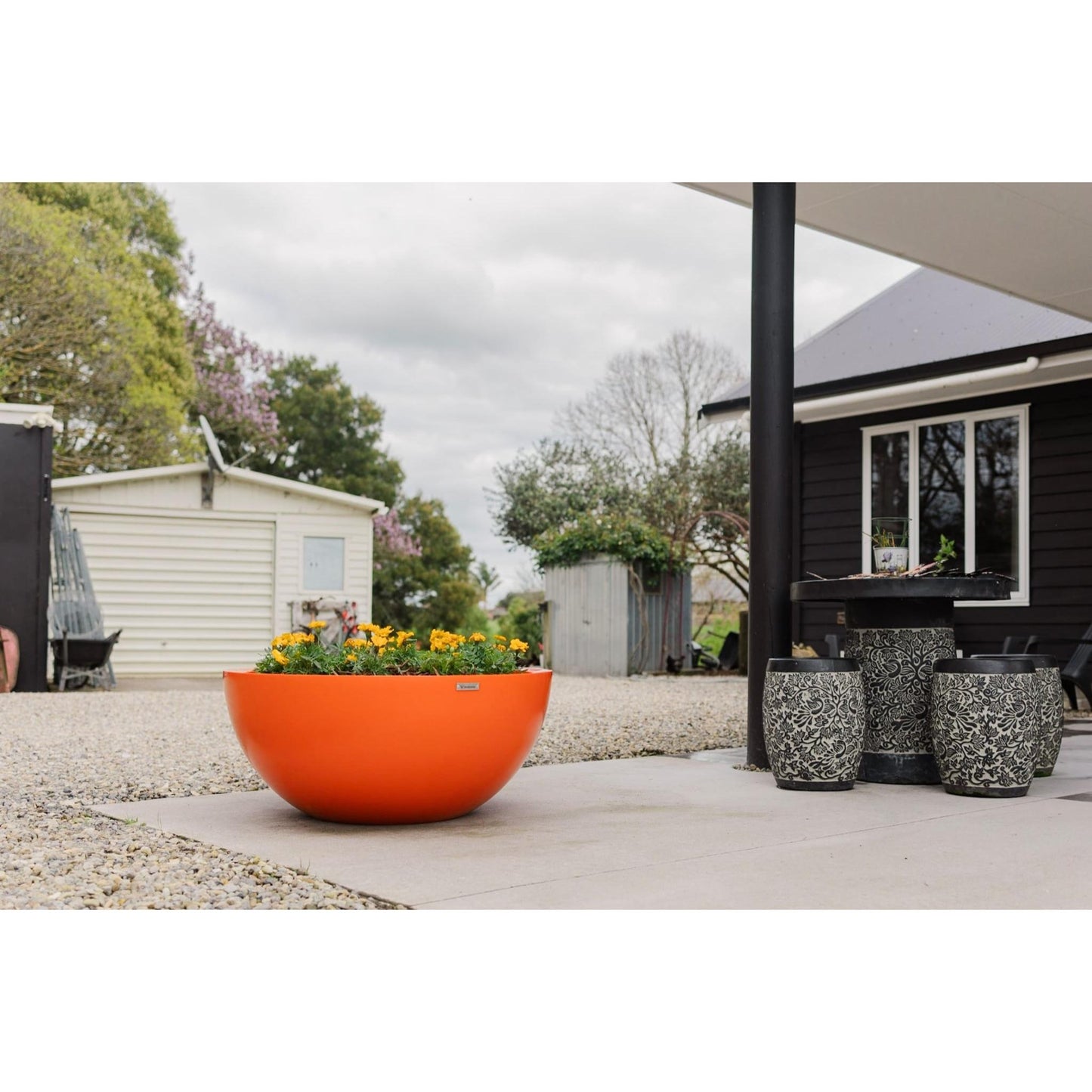 A large orange Modscene bowl planter pot on a concrete patio. The planter pot has Marigolds in it.