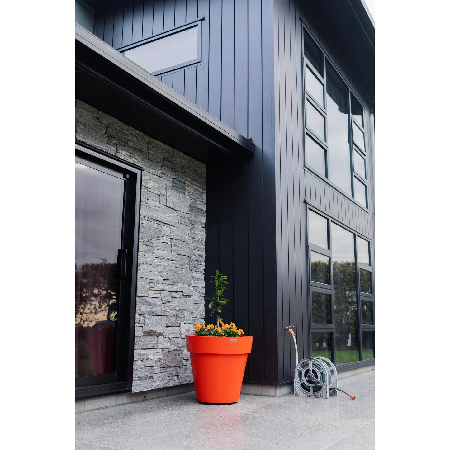 A large orange planter pot in front of a house wall. The pot is planted with a citrus tree and some flowers.