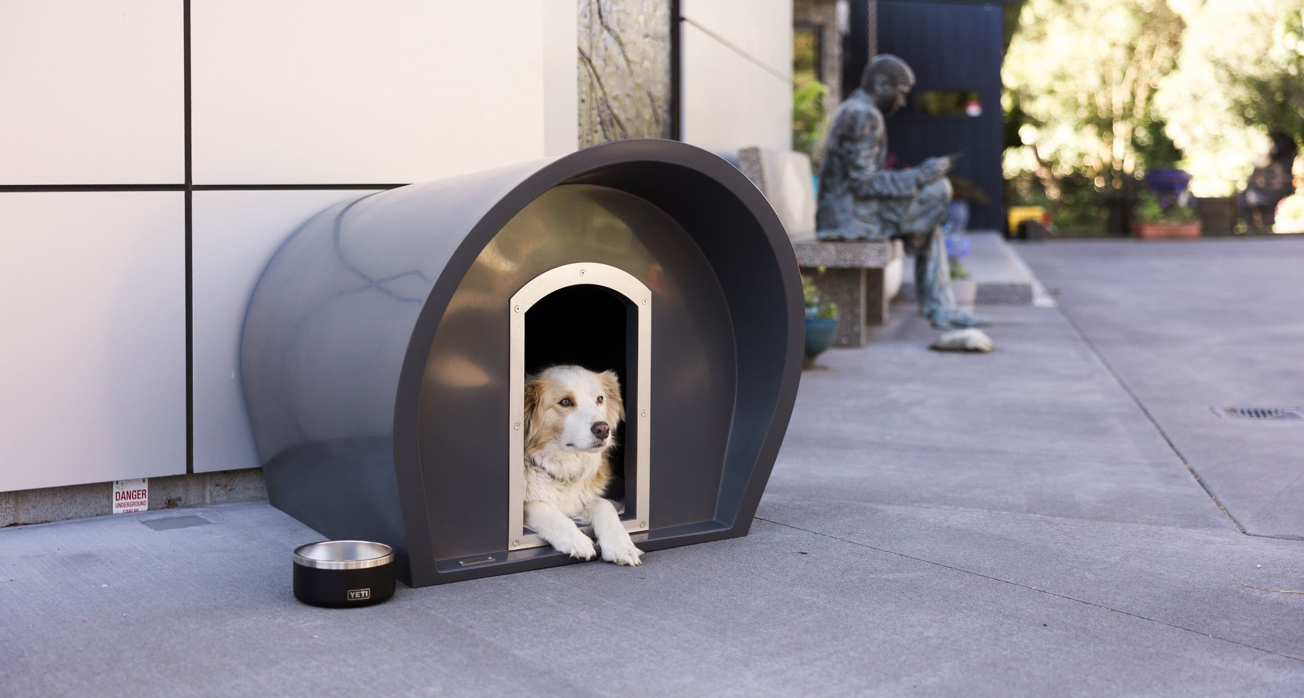 Dog peeking out from a modern doghouse on a concrete surface with a building in the background.