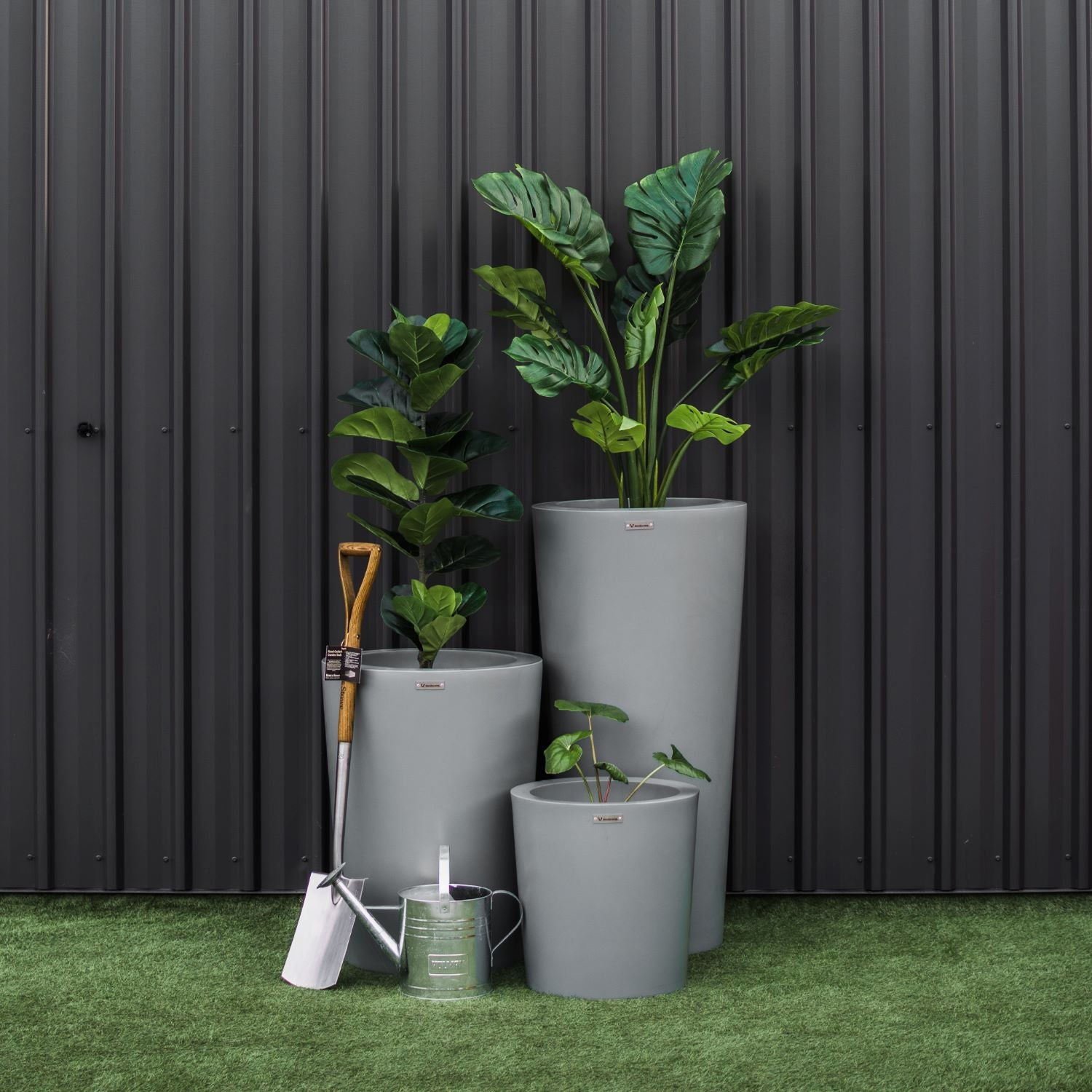 A cluster of grey Modscene planter pots. The pots have greenery in them and are in front of a black wall.