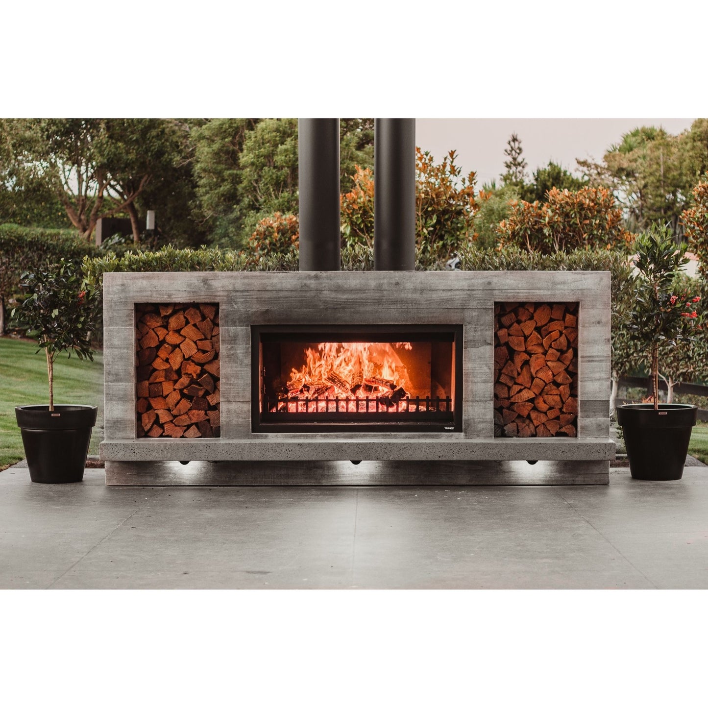 Large black Modscene planters by an outdoor fireplace. The fireplace is a Trendz Outdoors fireplace.