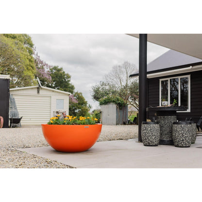 A large orange Modscene bowl planter pot on a concrete patio. The planter pot has Marigolds in it. 