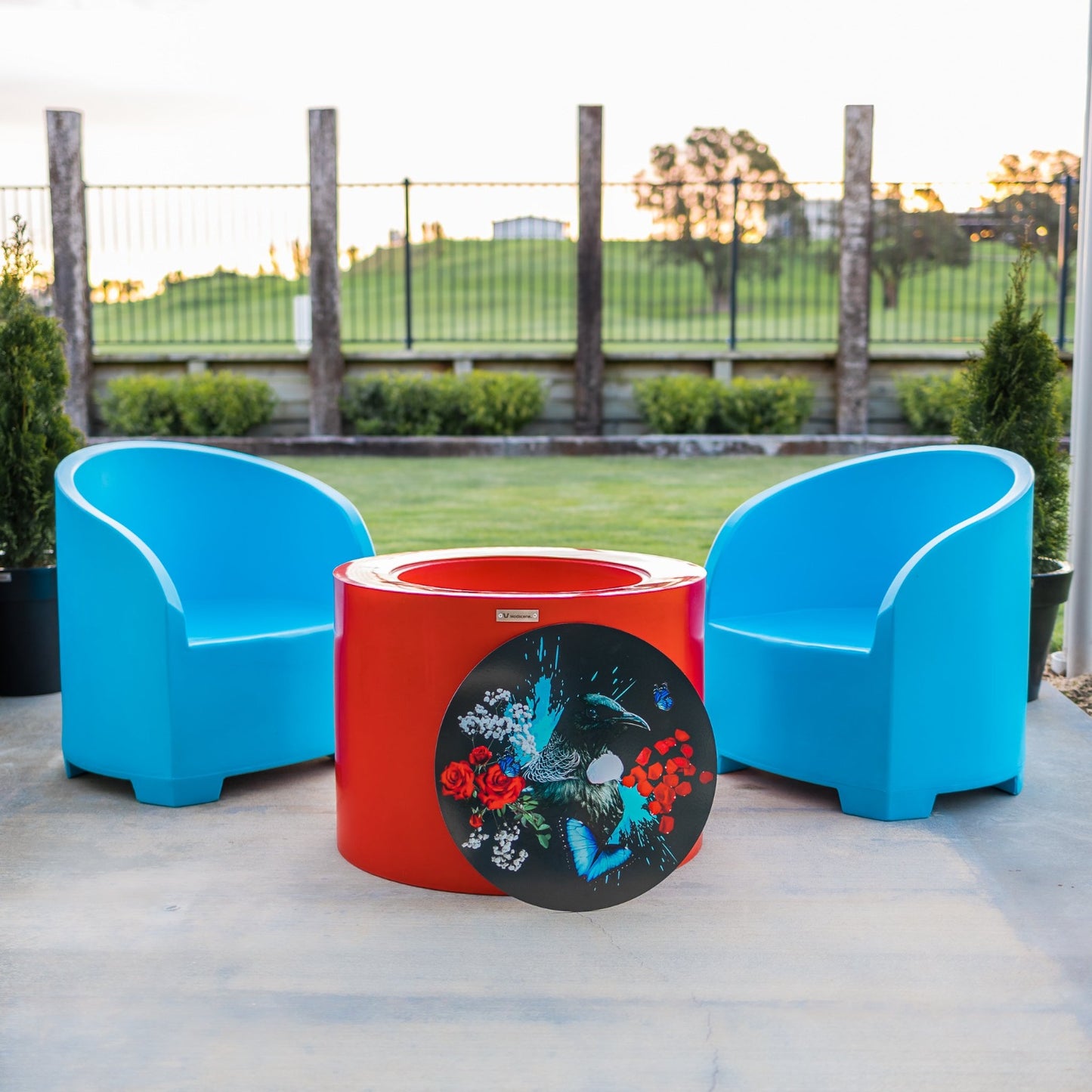 Two blue outdoor chairs and a red cylinder table with a Tui tabletop.